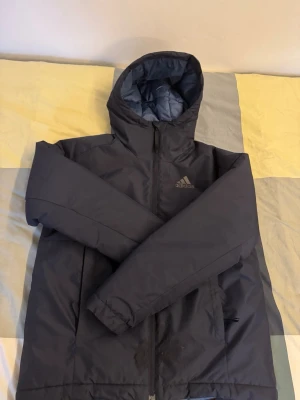 Mörkblå pufferjacka från Adidas - Säljer en mörkblå pufferjacka från Adidas med huva och dragkedja framtill. Jackan har en enkel och sportig design med diskret Adidas-logga på bröstet. Perfekt för kalla dagar och riktigt skön att bära.
