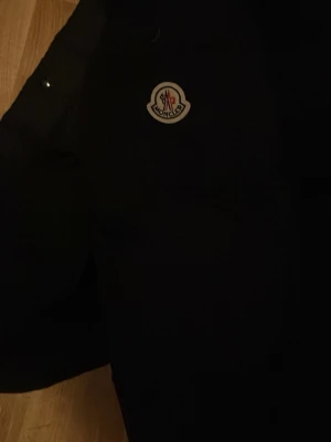 Svart Moncler dunjacka - Svart dunjacka från Moncler med klassisk logga på bröstet och coolt serietidningsmotiv på insidan. Jackan har tryckknappar och quiltad design som ger extra värme. Perfekt för dig som vill ha en snygg och varm jacka med exklusiv känsla.
