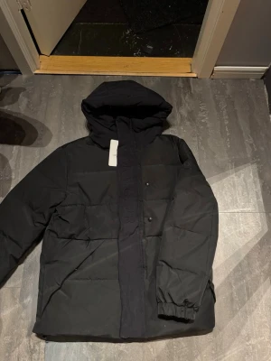 Moncler jacka - Säljer en svart dunjacka från Moncler med huva och diskret logga på ärmen. Jackan har en stilren look med broderad Moncler-text på framsidan, elastiska muddar och dragkedja med tryckknappar. Perfekt för kalla dagar när du vill hålla stilen.