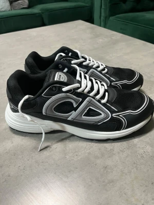 Svarta Dior sneakers med silverdetaljer - Säljer ett par svarta Dior sneakers med vita och silverfärgade detaljer. Skorna har en chunky vit sula, snörning och stora CD-loggor på sidorna och hälen. Ovandelen är i mesh och syntet med coola kontrastlinjer. Perfekta för dig som gillar statement pieces.