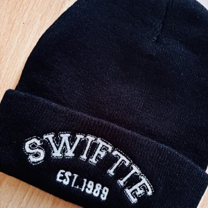 Svart stickad mössa med Swiftie-tryck! - Svart stickad mössa med "Swiftie est. 1989" broderat fram i Onesize. Oanvänd. 