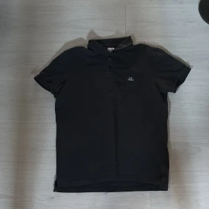 Mörkblå piké från C.P. Company - Mörkblå pikétröja från C.P. Company med klassisk krage och korta ärmar. Tröjan har broderad logga på bröstet och knappar framtill. Materialet är mjukt och känns som bomull, perfekt för en clean och stilren look.