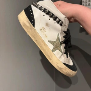 Golden Goose Mid Star sneakers med nitar - Säljer ett par Golden Goose Mid Star sneakers i vitt och svart skinn med coola silverfärgade nitar längs sidan och glittrig stjärndetalj. Skorna har svarta snören, beige sula och rund tå. Perfekt för dig som gillar edgy och unik stil. Skorna har ej dustbag eller box till har inget äktehetsbevis därav priset men kan nästan garantera att det är äkta då det har allt i sig som äkta golden goose ska ha. Det har lite defekter men funkar fint 