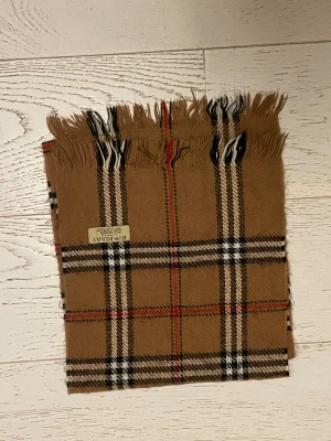 Burberry rutig halsduk i kashmir - Snygg halsduk från Burberry i klassiskt rutigt mönster med beige, svart, vit och röda detaljer. Gjord i 100% kashmir, supermjuk och lyxig. Fransar i ändarna och ikonisk Burberry-tag. Perfekt accessoar för att lyfta din stil.