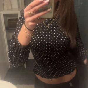 Svart prickig blus med puffärm - Säljer en svart blus med vita prickar och puffiga långärmar. Toppen har en croppad passform och rund halsringning, vilket ger en trendig och ungdomlig look. Perfekt för dig som gillar mönster och vill sticka ut lite extra.