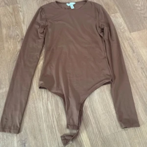 Brun långärmad bodysuit från H&M - Säljer en stilren brun bodysuit från H&M i storlek XS. Modellen har långa ärmar och rund halsringning, med en tight passform som sitter snyggt mot kroppen. Materialet är mjukt och stretchigt, perfekt för att skapa en clean och enkel look.