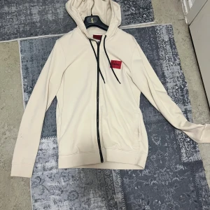 Beige hoodie från Hugo med dragkedja - Snygg beige hoodie från Hugo med svart dragkedja och snörning i huvan. Rött Hugo-märke på bröstet ger en clean och modern vibe. Tröjan har långa ärmar och är tillverkad i mjuk bomull, perfekt för chill dagar. Huvan är justerbar med svarta snören.