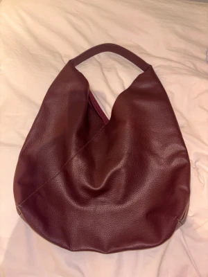 Stor vinröd axelväska - Säljer en stor och mjuk axelväska i vinrött skinn med enkel, stilren design. Väskan har ett brett handtag och en praktisk innerficka med dragkedja. Perfekt för dig som gillar minimalistisk look och vill ha plats för allt du behöver.