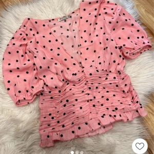 Rosa prickig blus från Stradivarius - Supersöt rosa blus från Stradivarius med svarta prickar, puffiga korta ärmar och v-ringad hals. Blusen har rynkade detaljer över midjan och en liten volangkant nedtill. Perfekt för dig som gillar färg och mönster!