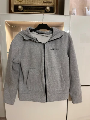 Grå hoodiejacka från Sail Racing - Grå hoodiejacka från Sail Racing med svart dragkedja och två stora fickor framtill. Insidan är fodrad med mjuk fleece för extra komfort. Jackan har en stilren look med logga på bröstet och ribbade detaljer vid kragen. Perfekt för chill dagar.