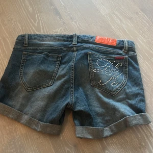 Blå jeansshorts från Dressed to Kill - Snygga blå jeansshorts från Dressed to Kill med uppvikta ben och klassisk femficksdesign. Ena bakfickan har coola strassdetaljer som ger extra edge. Passar perfekt till sommarens alla häng och har en normal passform med knapp och dragkedja framtill. Denna finns inte kvar längre så passa på att köpa!