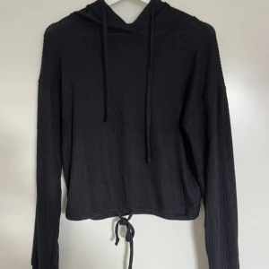 Svart ribbad hoodie från Gina Tricot - Svart hoodie från Gina Tricot i storlek S. Tröjan är ribbad, har långa ärmar, huva med snörning och en knytning nedtill som en extra detalj. Perfekt för dig som gillar en enkel men bekväm look.