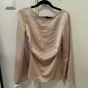 Champagnefärgad blus från Boohoo - Säljer en stilren blus från Boohoo i champagnefärgad satin. Blusen har lång ärm, rundad halsringning och ett lätt insytt midjeparti som ger en snygg siluett. Materialet är glansigt och mjukt, perfekt för dig som gillar en elegant vibe.