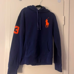 Marinblå hoodie från Polo Ralph Lauren - Snygg marinblå hoodie från Polo Ralph Lauren med orange logga på bröstet och en stor orange siffra 3 på ärmen. Tröjan har huva med snörning och en klassisk känguruficka framtill. Perfekt för dig som gillar sportig och stilren streetwear.