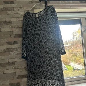 Svartvit mönstrad klänning från LMTX - Svartvit långklänning från LMTX med grafiskt rutigt mönster och kontrasterande kantdetaljer vid ärmslut och nederkant. Klänningen har rund hals och långa ärmar, tillverkad i mjukt och lätt material som ger en skön känsla. Perfekt för dig som gillar stilrena och unika prints.