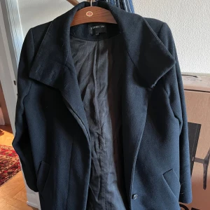 Svart kappa från STOCKH LM, stl 40 - Svart kappa från STOCKH LM i klassisk rak modell med bred krage och diskreta sidofickor. Kappan är helsvart och har knappar vid ärmslut och framtill. Materialet är ullblandning som ger en snygg och stilren look. Perfekt för dig som gillar minimalistisk stil.