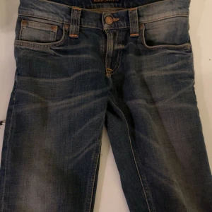 Blå jeans från Nudie Jeans Co - Snygga blå jeans från Nudie Jeans Co med klassisk femficksdesign och orangea sömmar. Jeansen har en mörk tvätt med slitningar och Nudie-logga på bakfickan. Passformen är rak och materialet är robust denim. Perfekt för dig som gillar streetwear och tidlös stil.