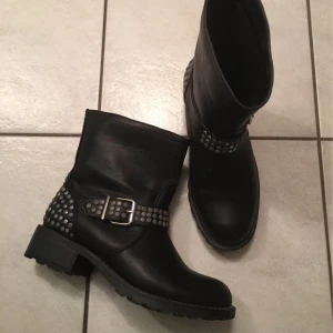 Svarta boots med nitar och spänne - Snygga svarta boots i skinn med dekorativa nitar och ett spänne runt ankeln. Perfekta för en tuff look med en robust sula och låg klack.