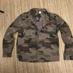 Camouflage overshirt  - Säljer en snygg camouflage overshirt i storlek M. Den har ett coolt broderat mönster på ryggen med text i lila. Perfekt för höst och vår, med långa ärmar och knappar framtill. Passar både till vardags och fest!