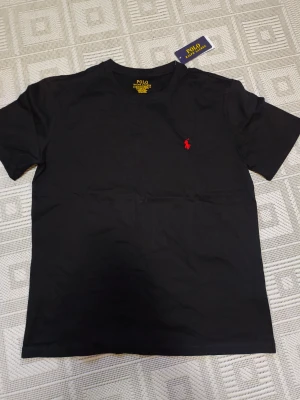 Svart t-shirt PRL - Svart t-shirt från PRL med klassisk röd broderad logga på bröstet. Modellen är slim fit och har rund hals samt korta ärmar. Tillverkad i mjuk bomull för skön känsla. Perfekt för dig som gillar stilrena och ikoniska plagg.