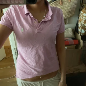 Rosa pikétröja från Ralph Lauren - Säljer en ljusrosa pikétröja från Ralph Lauren med klassisk krage och korta ärmar. Tröjan har två knappar vid halsen och den ikoniska broderade loggan på bröstet. Perfekt för en chill och snygg look.