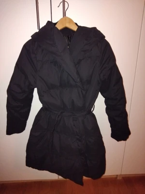 Svart lång omlott dunjacka med bälte - Svart omlott dunjacka med mellanlång passform och bälte i midjan. Jackan har huva och långa ärmar, perfekt för kalla dagar. Stilren och enkel design som håller dig varm hela vintern.