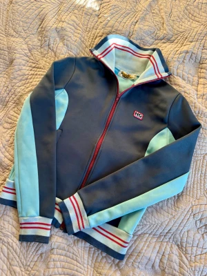 Hysteric Glamour zip - Snygg ziptröja från Hysteric Glamour i mörkblå och ljusblå färg med röda detaljer. Tröjan har lång ärm, högt krage och HG-logga på bröstet. Ränder i rött och vitt på muddar och krage, samt hel dragkedja framtill. Perfekt för dig som gillar retrostil. Strlk S/XS