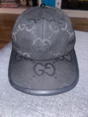 Svart Gucci keps med GG-mönster - Snygg svart keps från Gucci med det klassiska GG-mönstret över hela kepsen. Kepsen har böjd skärm, justerbar rem bak i svart och är tillverkad i slitstark bomull. Perfekt för dig som vill ha en lyxig streetstyle-accessoar.