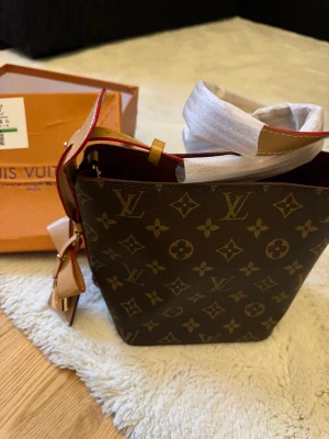 Monogram axelväska - Snygg axelväska med klassiskt monogrammönster i brunt och beige. Väskan har en fyrkantig form, beige axelrem med röda detaljer och guldfärgade metalldetaljer. Perfekt storlek för det viktigaste och en ikonisk look.