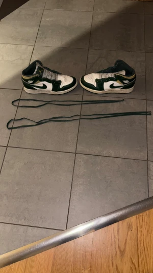 Nike Air Jordan 1 Mid vit/grön/gul - Nike Air Jordan 1 Mid sneakers i vit, mörkgrön och gul färg. Klassisk high-top siluett, perforerad tå och gröna detaljer på sidorna samt gul logga på plösen. Skorna har en platt sula och är tillverkade i skinn och syntetmaterial. Box medföljer. Pris kan diskuteras.