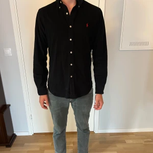 Svart skjorta från Ralph Lauren - Klassisk svart skjorta från Ralph Lauren med knappar hela vägen och röd broderad logga på bröstet. Skjortan har lång ärm och normal passform, perfekt till många olika tillfällen.