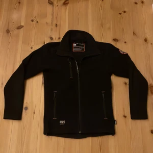 Svart fleecejacka Helly Hansen XS - Svart fleecejacka från Helly Hansen Workwear i storlek XS. Jackan har hel dragkedja, hög krage och tre fickor med dragkedja. Snyggt HH-märke på ärmen och detaljer i orange. Perfekt för kyliga dagar och outdoor. Relativt använd men fortfarande i bra skick
