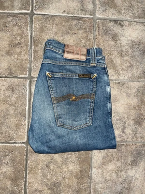 Nudie jeans  - Snygga blå jeans från Nudie Jeans med klassisk femficksdesign och gul broderad detalj på bakfickorna. Modellen är slim med smal passform. OBS vet ej om hålen är design heller defekt! Längd 103cm. JAG SKICKAR EJ FLER BILDER NÄR JAG BÄR JEANSEN! för storleksguide hänvisar jag till måtten och för bättre inblick i passformen rekommenderar jag att googla på modellnamnet. :) 50 