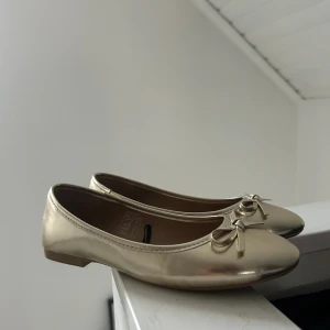 Guldiga ballerinaskor med rosett - Snygga ballerinaskor i metallic guld från Claudia Ghizzani. Skorna har rund tå, platt sula och en söt rosettdetalj framtill. Tillverkade i syntetmaterial med en mjuk innersula för extra komfort. Perfekt för dig som vill ha en glammig touch på outfiten.