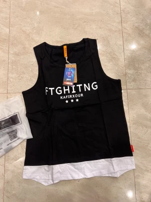 Svart och vit linnetopp med RUN-tryck - Cool svart linnetopp från Kafirxour med bred vit kant längst ner och stora vita tryck på både fram- och baksida. Framsidan har texten 'FTGHITNG KAFIRXOUR' och tre stjärnor, baksidan har ett stort 'RUN'. Perfekt för dig som gillar sportig streetstyle.