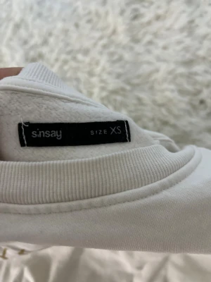 Vit sweatshirt med guldtryck Sinsay XS - Vit sweatshirt från Sinsay i storlek XS med snyggt tryck i guld och svart på framsidan. Tröjan har rund hals, långa ärmar med puff och resår vid ärmslut och nederkant för en cool och avslappnad look. Perfekt för dig som gillar statement-plagg.