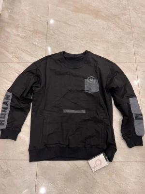 Svart sweatshirt med grå detaljer HUMAN - Svart sweatshirt från HUMAN med grå ficka på bröstet och grå paneler på ärmarna. Tryckt text på ena ärmen och coolt tryck vid bröstfickan. Rund hals, ribbade muddar och två sidofickor. Perfekt för dig som gillar streetwear och snygga kontraster.