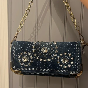 Blå axelväska med pärlor och kedja - Snygg axelväska i mörkblått med silverfärgade detaljer och pärlor på framsidan. Väskan har ett flätat mönster på baksidan och en guldfärgad kedja som axelrem. Insidan har flera fack och dragkedja. Perfekt för dig som gillar Y2k stilen!