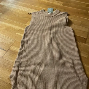 Beige ribbad stickad klänning Aware - Ribbstickad beige klänning från Aware med hög krage och ärmlös design. Klänningen har en rak, loose passform och är tillverkad i ett mjukt stickat material. Perfekt för lager-på-lager och chill vibes.