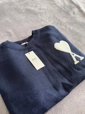 Ami Paris tröja  - Snygg marinblå stickad tröja från Ami Paris med rund hals och ett vitt hjärta med A på bröstet. Tröjan har ribbade muddar och är tillverkad i mjuk ull som känns riktigt skön mot huden. Perfekt för dig som gillar stilrena detaljer och vill ha något som sticker ut.