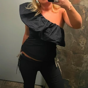 Svart oneshoulder volangtop - Säljer en svart oneshoulder topp med stor volang över axeln. Toppen har en cool asymmetrisk design och sitter tight över kroppen. 