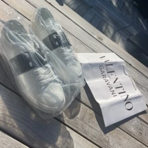 Valentino Garavani vita sneakers - Snygga vita sneakers från Valentino Garavani med bred svart detalj över sidan och klassisk snörning. Skorna är inte äkta-Skorna har rund tå och platt sula, tillverkade i skinn för en lyxig känsla. Perfekta för dig som vill ha en clean och stilren look med designer-vibe.