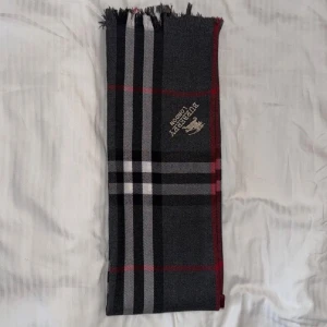 Rutig halsduk från Burberry London - Snygg halsduk från Burberry London med klassiskt rutigt mönster i mörkgrått, svart, vitt och röda detaljer. Broderad logga och fransad kant. Perfekt accessoar för att lyfta din outfit med lite lyxig London-vibe.