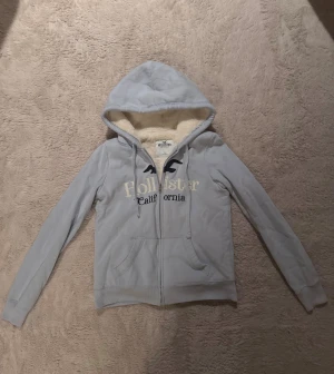 Ljusblå hoodiejacka från Hollister - Mysig ljusblå hoodiejacka från Hollister med broderad logga och text på bröstet. Jackan har dragkedja, snörning i huvan och är fodrad med fluffigt, krämfärgat teddyfoder. Perfekt för kyliga dagar och riktigt skön att ha på sig.