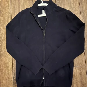 Mörkblå kofta med dragkedja från Bläck - Svart kofta från Bläck i storlek M. Tröjan har en ribbad krage och muddar, hel dragkedja framtill och är långärmad. Materialet är mjukt och stickat, perfekt för kyliga dagar. Enkel och stilren design som funkar till många olika looks.