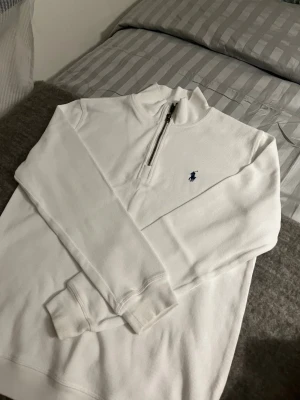 Vit half zip tröja från Polo Ralph Lauren - Säljer en clean vit långärmad tröja från Polo Ralph Lauren med blå logga på bröstet. Tröjan har en snygg half zip-dragkedja och ribbade muddar. Perfekt till lager på lager och funkar året runt.