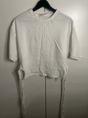 Vit croppad topp med knyt från Nelly - Säljer en vit croppad topp från Nelly med korta ärmar och rund hals. Toppen har långa knytband i sidorna som ger en snygg detalj och kan justeras för att passa din stil. Perfekt till jeans eller kjol för en clean look. ❤️