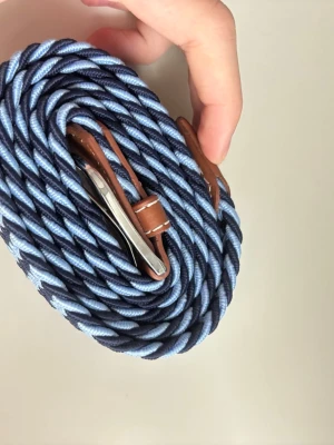 Flätat blått och brunt bälte - Säljer ett snyggt flätat bälte i marinblått och ljusblått med detaljer i brunt skinn och silverfärgat spänne. Perfekt accessoar för att lyfta din outfit med en cool och modern touch.