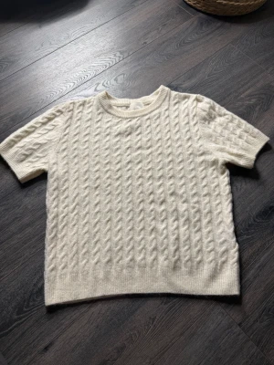 Vit kabelstickad kortärmad tröja - Använd 1 gång. Säljer en vit kabelstickad tröja med korta ärmar och rund hals. Tröjan har ett klassiskt flätmönster över hela och är gjord i ett mjukt, stickat material. Perfekt för lager på lager under höst och vår.