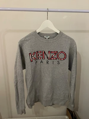 Grå tröja från Kenzo Paris - Säljer en grå sweatshirt från Kenzo Paris i storlek M. Tröjan har ett stort Kenzo-tryck i rött och vitt framtill och är tillverkad i mjuk bomull. Klassisk rund halsringning och långa ärmar gör den perfekt för chill dagar.